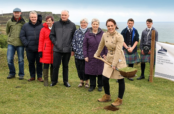 Members of the community and Flodigarry Township Trust look on as local MSP Kate Forbes cuts the first turf at the ceremony .